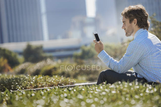 Joven sentado en el banco del parque en la ciudad y usando el teléfono móvil . - foto de stock
