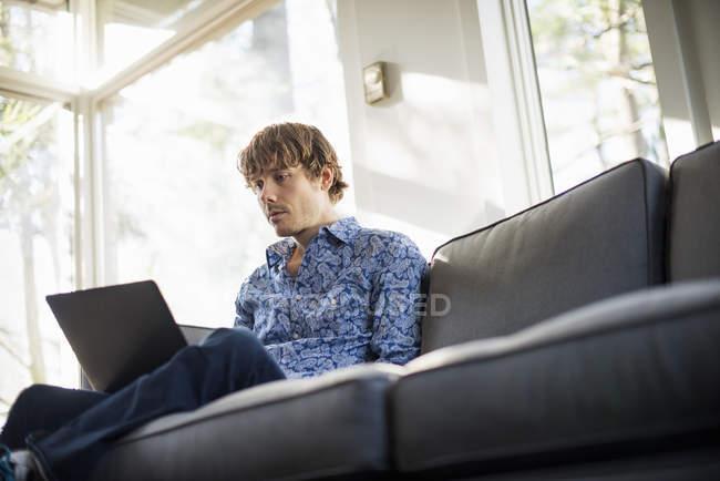 Vista de ángulo bajo del hombre sentado en el sofá y mirando a la computadora portátil . - foto de stock