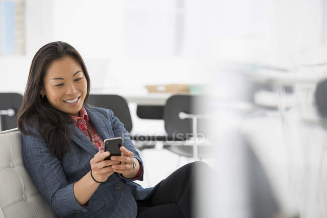 Femme vérifiant smartphone sur chaise confortable dans l'intérieur de bureau moderne . — Photo de stock