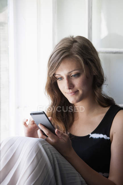 Молода жінка сидить біля вікна і використовує смартфон . — стокове фото