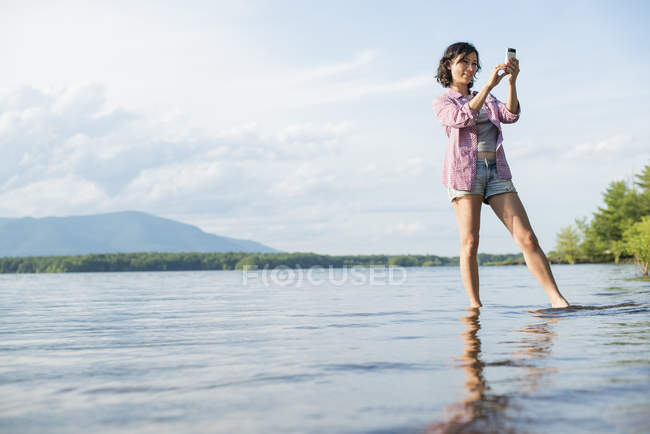 Жінка стоїть у воді озера і фотографує зі смартфоном . — стокове фото