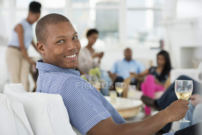 Молодой человек с бокалом вина сидит в кресле с людьми, веселящимися на заднем плане . — стоковое фото