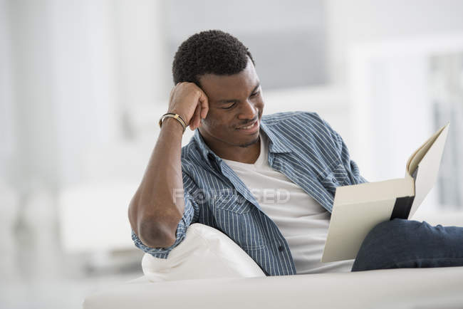 Junger Mann sitzt und liest Buch im hellen weißen Raum. — Stockfoto