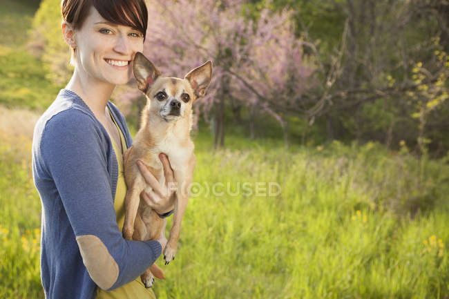 Giovane donna che tiene e abbraccia il cane chihuahua sul campo di erba nel parco
. — Foto stock
