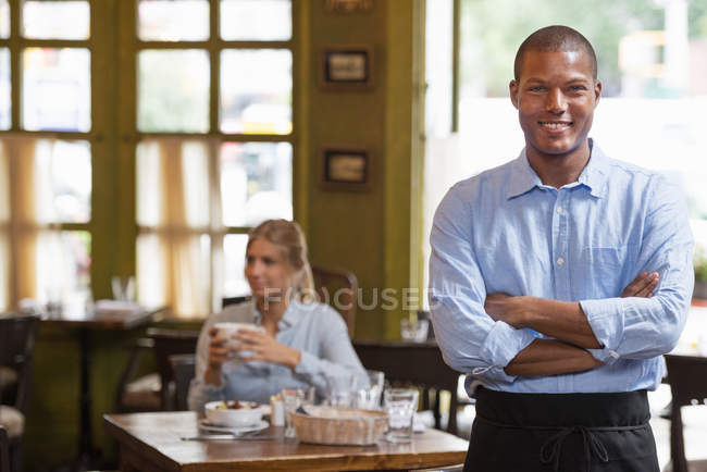 Мужчина-официант, стоящий со сложенными руками в интерьере кафе с женщиной, держащей кофе на заднем плане . — стоковое фото