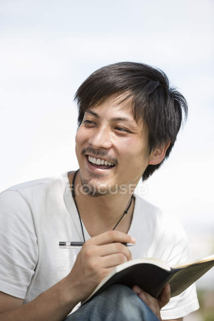 Young man laughing, holding pen and notebook and looking away outdoors. — Stock Photo