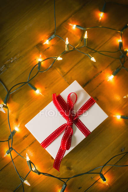 Caixa de presente amarrada com fita vermelha e luzes de fadas no chão de madeira . — Fotografia de Stock