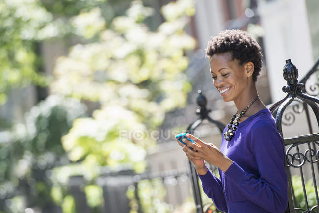 Mulher de vestido roxo apoiando-se na cerca na rua e usando smartphone . — Fotografia de Stock