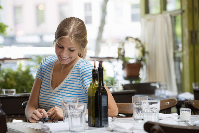 Frau im gestreiften T-Shirt sitzt am Cafétisch und checkt Smartphone. — Stockfoto
