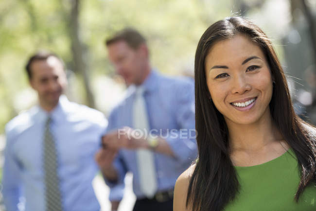 Mid adult woman smiling and looking in camera with businessmen talking in background. — Stock Photo
