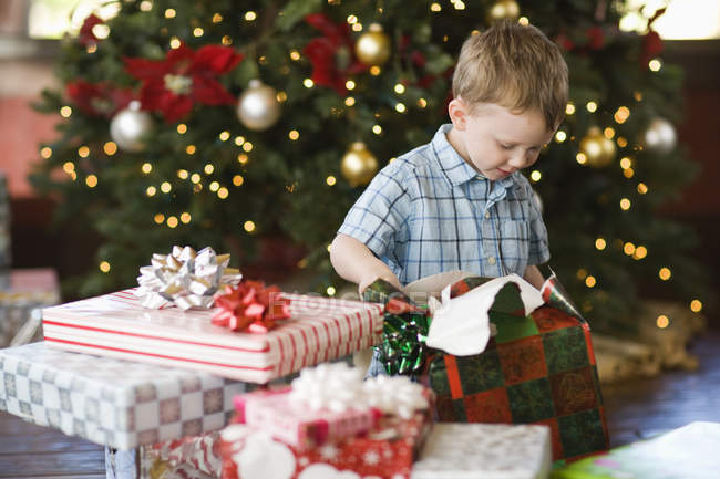 Niño de edad elemental sentado y desenvolviendo regalos bajo el árbol de Navidad . - foto de stock