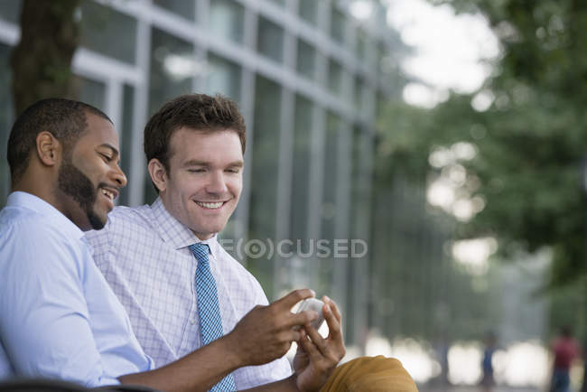 Two businessmen looking at smartphone while sitting on bench in downtown. — Stock Photo