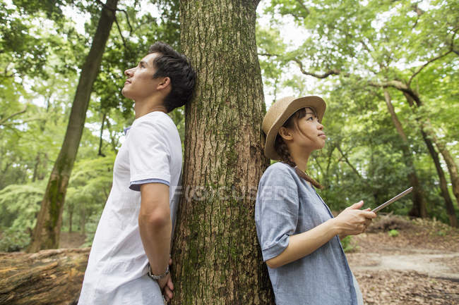 Японская пара опирается на ствол дерева в лесу, женщина держит цифровой планшет . — стоковое фото