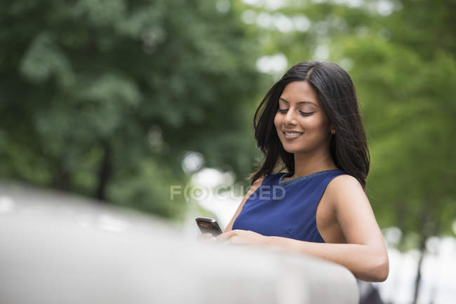 Frau mit langen schwarzen Haaren in blauem Kleid benutzt Smartphone im Park. — Stockfoto