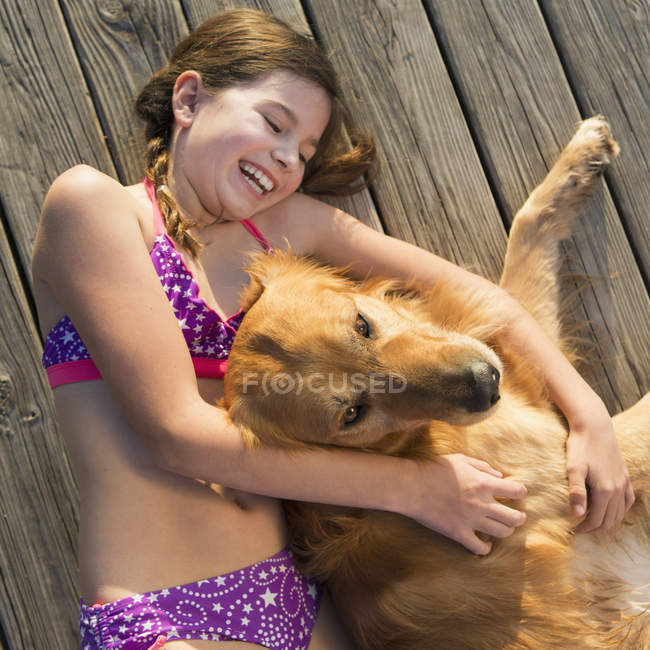 Pre-adolescent girl in swimwear with golden retriever dog lying on wooden pier. — Stock Photo