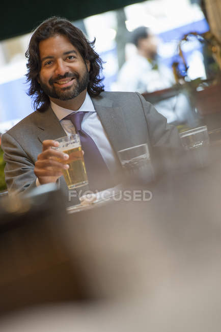 Homem hispânico sentado à mesa em bar interior e segurando vidro de cerveja e olhando para a câmera . — Fotografia de Stock