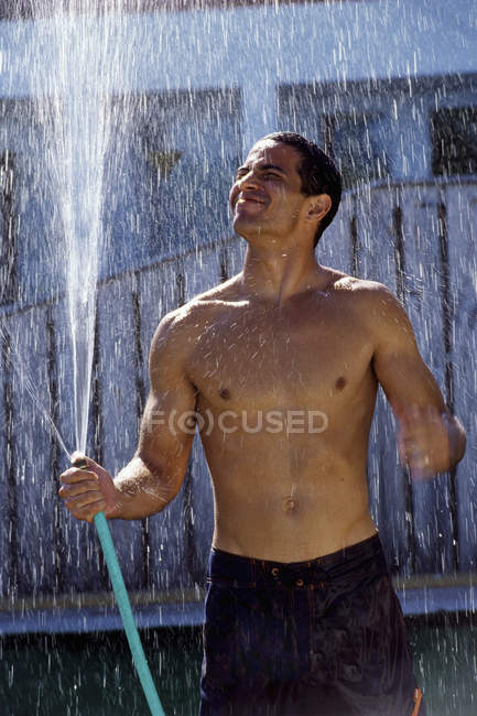Mann in kurzen Hosen mit nackter Brust hält Gartenschlauch in der Hand und steht im Wasserstrahl. — Stockfoto