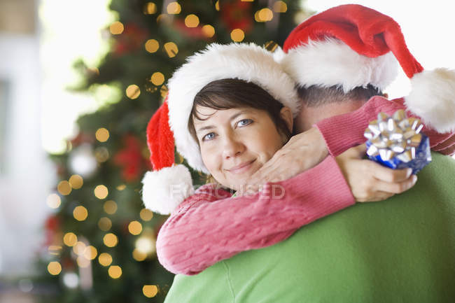 Couple wearing Santa hats hugging in front of Christmas tree. — Stock Photo