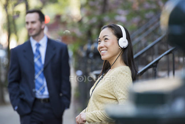 Donna che indossa cuffie musicali e uomo in giacca e cravatta camminare in background . — Foto stock