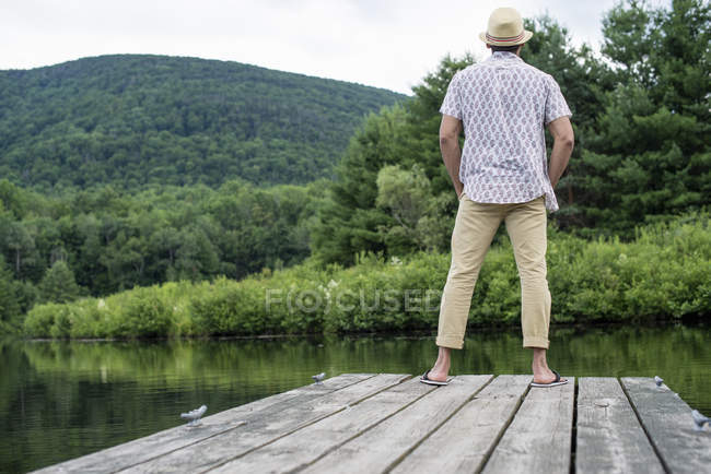 Rear view of man standing on wooden pier overlooking calm lake. — Stock Photo