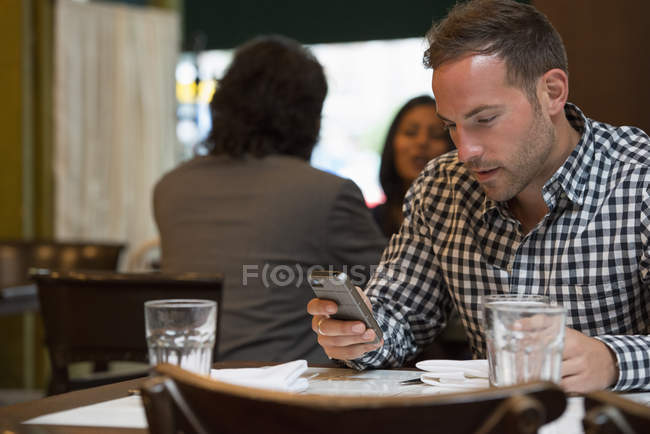 Man at cafe table checking phone with couple talking in background. — Stock Photo