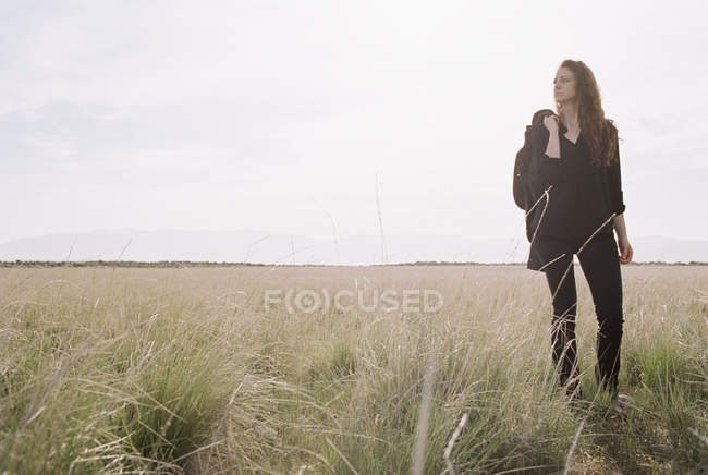 Donna che cammina attraverso il paesaggio erboso portando giacca e zaino . — Foto stock