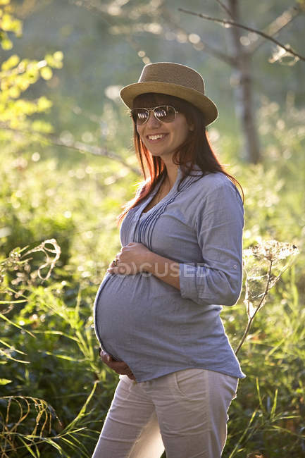 Femme enceinte en chapeau et lunettes de soleil avec les mains sur le ventre dans le jardin . — Photo de stock