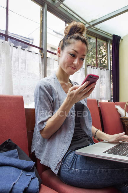 Mujer sonriendo mientras usa el teléfono inteligente y el portátil en sillas rojas en el interior . - foto de stock