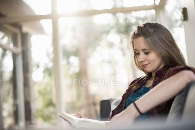 Sorrindo mulher sentada no sofá e lendo livro . — Fotografia de Stock