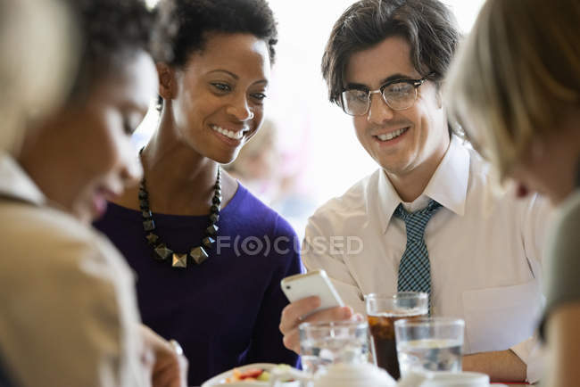 Homem e mulher compartilhando smartphone na mesa do restaurante com amigos . — Fotografia de Stock