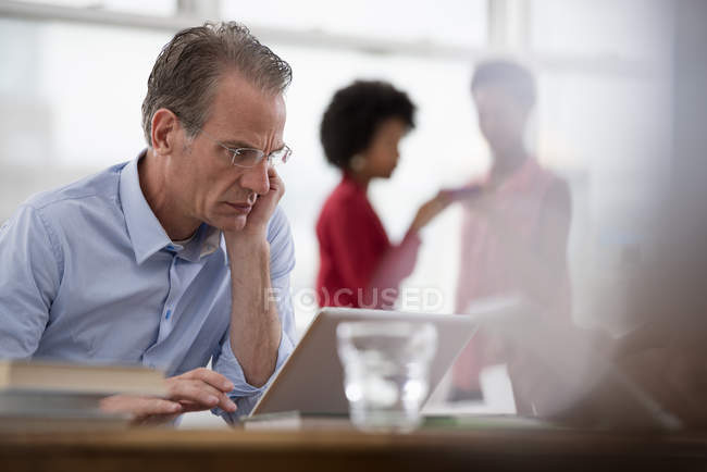 Зрілий чоловік використовує ноутбук на робочому місці з колегами на задньому плані . — стокове фото