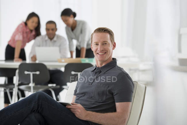 Man sitting on office armchair with colleagues using laptop in background. — Stock Photo