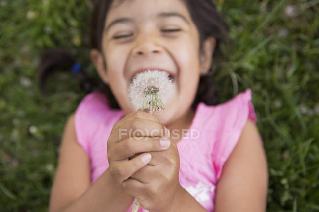 Elementary age girl lying on ground and holding dandelion seedhead, overhead view. — Stock Photo