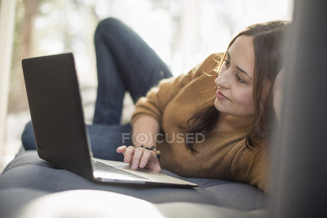 Весела молода жінка лежить на дивані і використовує ноутбук . — стокове фото