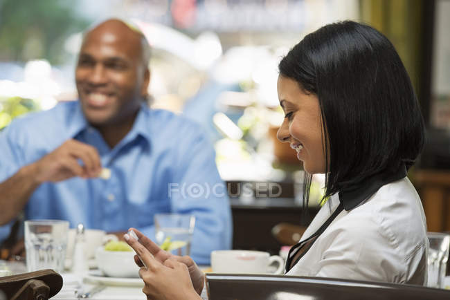 Жінка посміхається під час перевірки телефону за столиком кав'ярні з чоловіком, який їсть на задньому плані . — стокове фото