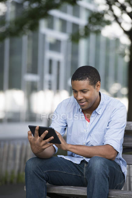 Afrikanisch-amerikanischer Mann nutzt digitales Tablet, während er auf Bank in der Stadt sitzt. — Stockfoto