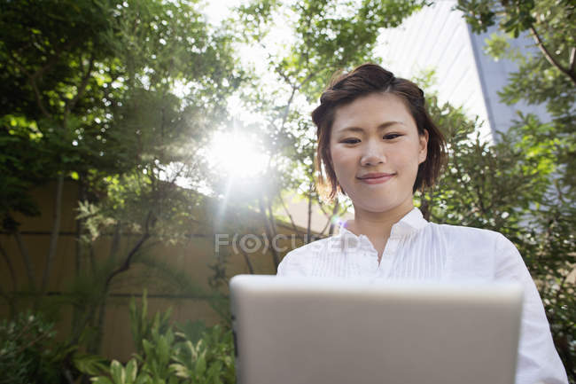 Jovem mulher japonesa usando laptop no jardim da cidade . — Fotografia de Stock