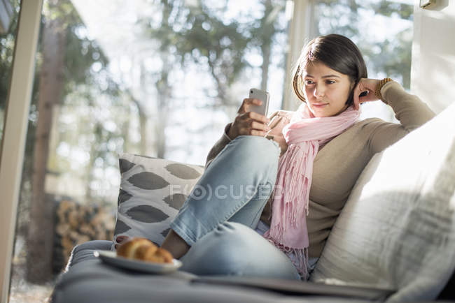 Mulher sentada no sofá e usando smartphone com placa com croissant na frente . — Fotografia de Stock