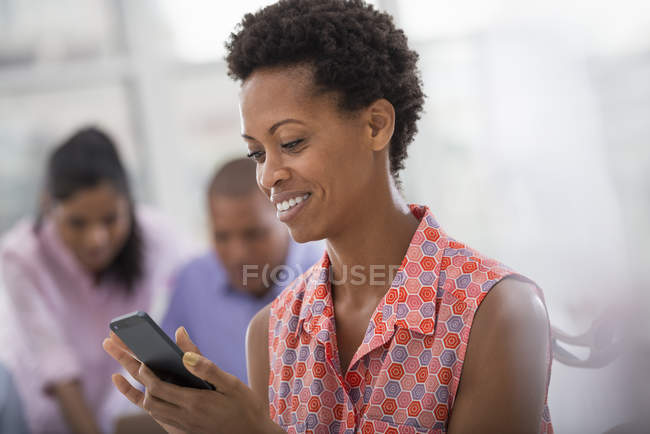 Smiling woman using smartphone with colleagues in background. — Stock Photo