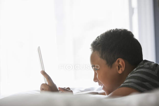 Vista laterale del ragazzo in età elementare utilizzando tablet computer sul letto . — Foto stock