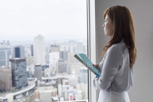 Vista lateral de la mujer de negocios sosteniendo archivos y mirando a través de la ventana en el edificio de oficinas . - foto de stock