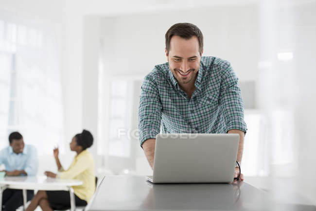 Mann stützt sich auf Tisch und benutzt Laptop mit Leuten, die im Büro sitzen und reden. — Stockfoto