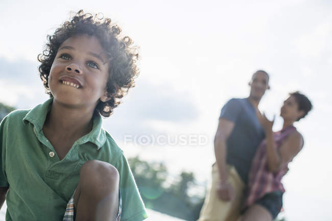 Elementary age boy sitting on sunny lake jetty with parents. — Stock Photo