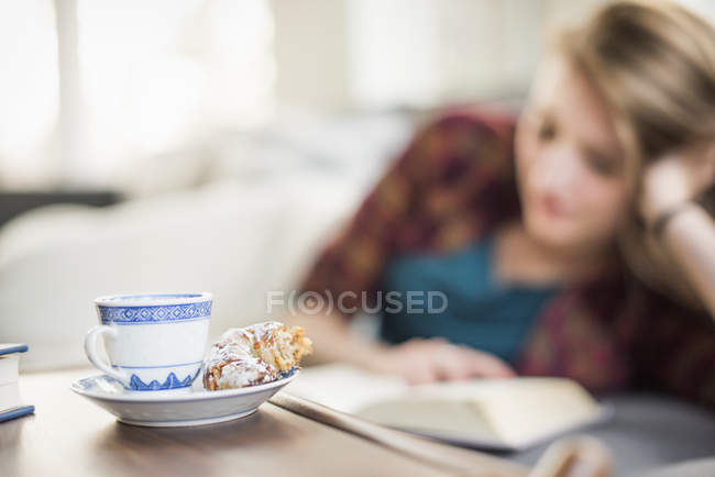 Tazza e piattino con croissant sul tavolo con libro di lettura donna sullo sfondo . — Foto stock