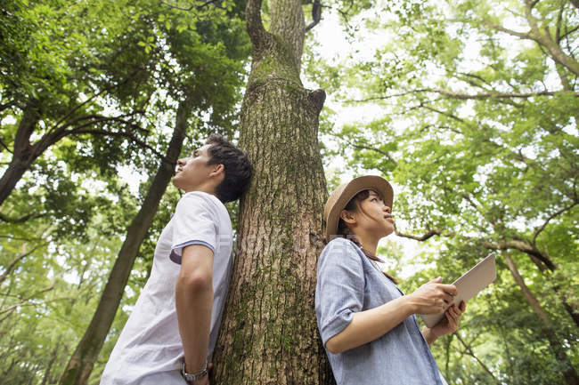 Japanisches Paar lehnt an Baumstamm im Wald, Frau hält digitales Tablet. — Stockfoto