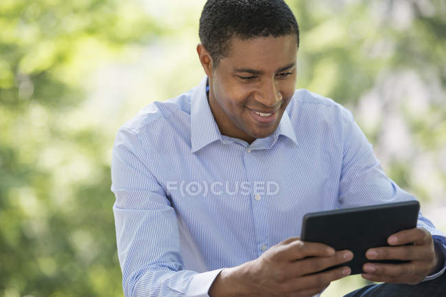 Uomo adulto mezzo utilizzando tablet digitale mentre seduto nel parco della città . — Foto stock