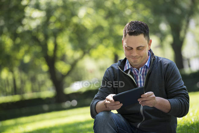 Hombre adulto medio usando tableta digital mientras está sentado en el parque de la ciudad . - foto de stock