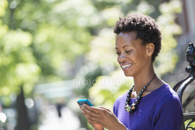 Frau mit Halskette checkt Smartphone und lächelt auf der Straße. — Stockfoto