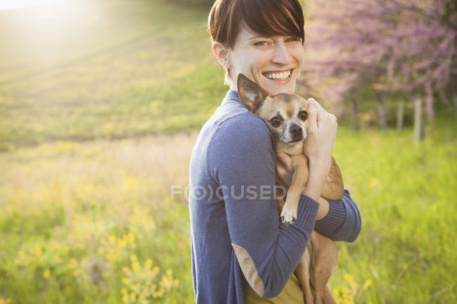 Giovane donna che tiene e abbraccia il cane chihuahua sul campo di erba nel parco . — Foto stock