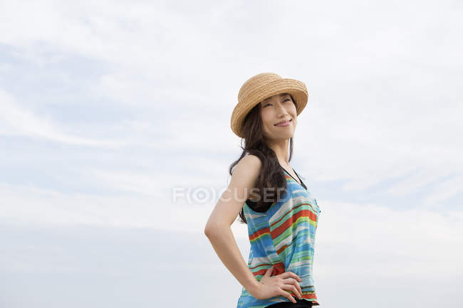 Cheerful Asian woman with straw hat standing outdoors against cloudy sky. — Stock Photo